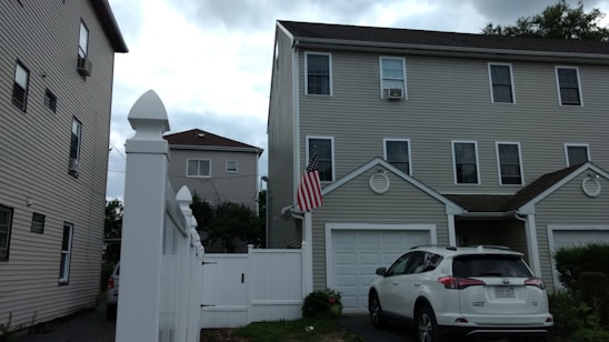 A residential area with a three-story house featuring multiple windows and a garage, alongside a white fence. An SUV is parked in front of the garage, and an American flag is mounted near the entrance. The sky is overcast with clouds.
