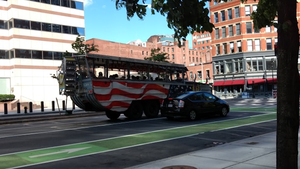 A street scene featuring an amphibious tour vehicle decorated with an American flag design parked on a multi-lane road. The vehicle is followed by a black car. The background includes tall brick buildings and a modern white building. Trees with green leaves line the sidewalk, partially shadowing the scene.