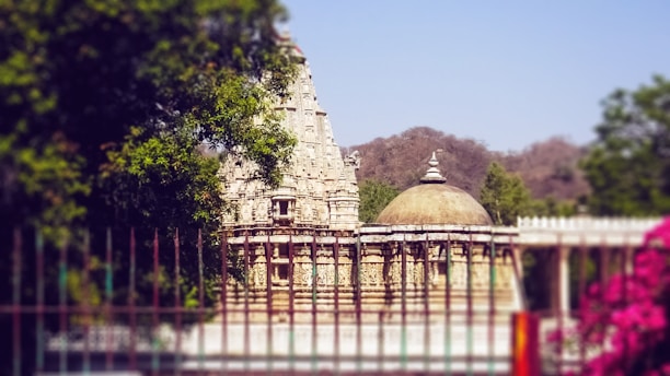 A historic temple with intricate stone carvings stands amidst lush greenery. The temple features a tall, ornate spire and a dome, encapsulated by trees and a garden fence. Pink flowers are in the foreground, while hills can be seen in the background under a clear blue sky.