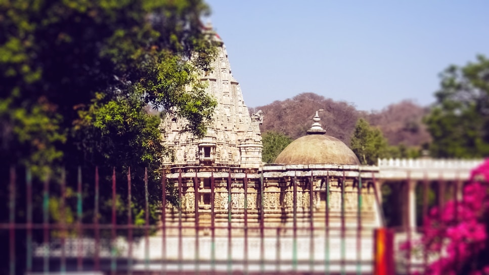 A historic temple with intricate stone carvings stands amidst lush greenery. The temple features a tall, ornate spire and a dome, encapsulated by trees and a garden fence. Pink flowers are in the foreground, while hills can be seen in the background under a clear blue sky.