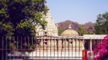 A historic temple with intricate stone carvings stands amidst lush greenery. The temple features a tall, ornate spire and a dome, encapsulated by trees and a garden fence. Pink flowers are in the foreground, while hills can be seen in the background under a clear blue sky.