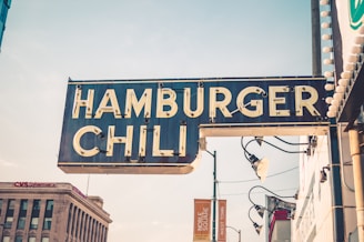 A vintage-style sign with the words 'Hamburger Chili' is prominently displayed. The sign is attached to a building and features retro lightbulbs around its edges. It's daytime with a slightly overcast sky, and a CVS Pharmacy building is visible in the background, alongside some streetlamps and additional signage.