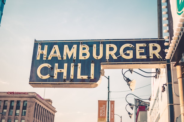 A vintage-style sign with the words 'Hamburger Chili' is prominently displayed. The sign is attached to a building and features retro lightbulbs around its edges. It's daytime with a slightly overcast sky, and a CVS Pharmacy building is visible in the background, alongside some streetlamps and additional signage.
