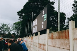 A large digital scoreboard at an outdoor sports venue displays a score of 2-2. Spectators are gathered, observing the event with interest. The scene is enclosed by a tall metal fence and surrounded by lush green trees, indicating a natural setting. Overcast skies add a muted tone to the environment.