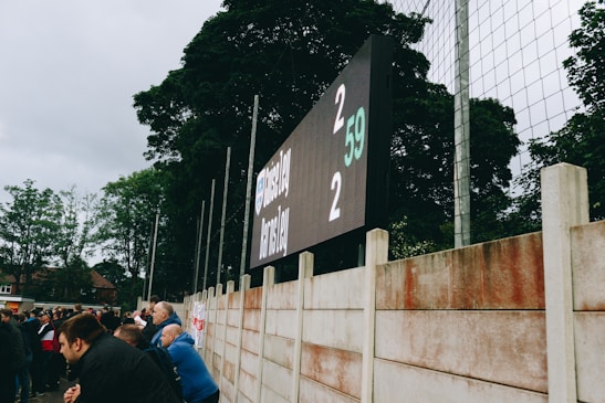 A large digital scoreboard at an outdoor sports venue displays a score of 2-2. Spectators are gathered, observing the event with interest. The scene is enclosed by a tall metal fence and surrounded by lush green trees, indicating a natural setting. Overcast skies add a muted tone to the environment.