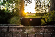 A portable Bluetooth speaker perched on a picnic blanket in a sunny park setting.