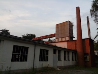 An industrial building with a long, rusty pipe running horizontally across the upper part. The structure features brick walls and tall brick chimneys, with a large metal installation on the roof. The building appears old and somewhat abandoned, surrounded by overgrown vegetation.