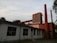 An industrial building with a long, rusty pipe running horizontally across the upper part. The structure features brick walls and tall brick chimneys, with a large metal installation on the roof. The building appears old and somewhat abandoned, surrounded by overgrown vegetation.