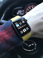 A person's wrist wearing a smartwatch with a yellow band, displaying a podcast app called 'THE DAILY' with a play button and a timestamp of 7:22. The background shows part of a car steering wheel with a distinctive logo.
