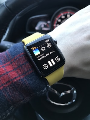 A person's wrist wearing a smartwatch with a yellow band, displaying a podcast app called 'THE DAILY' with a play button and a timestamp of 7:22. The background shows part of a car steering wheel with a distinctive logo.