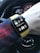 A person's wrist wearing a smartwatch with a yellow band, displaying a podcast app called 'THE DAILY' with a play button and a timestamp of 7:22. The background shows part of a car steering wheel with a distinctive logo.