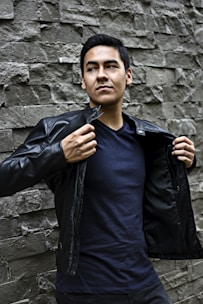 A model wearing a black leather jacket with edgy patches standing against a brick wall.
