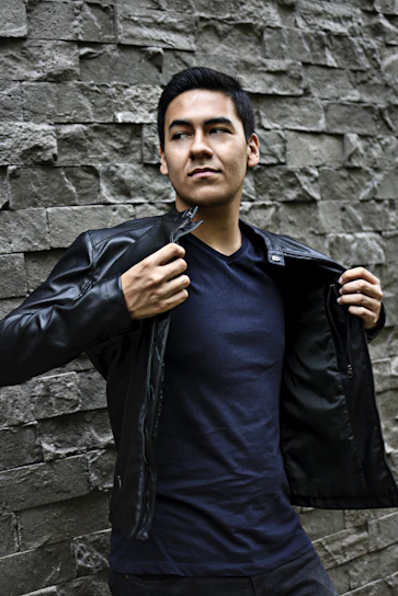 A stylish young man wearing a sleek black leather jacket and cream t-shirt, standing confidently against a minimalist brown backdrop.