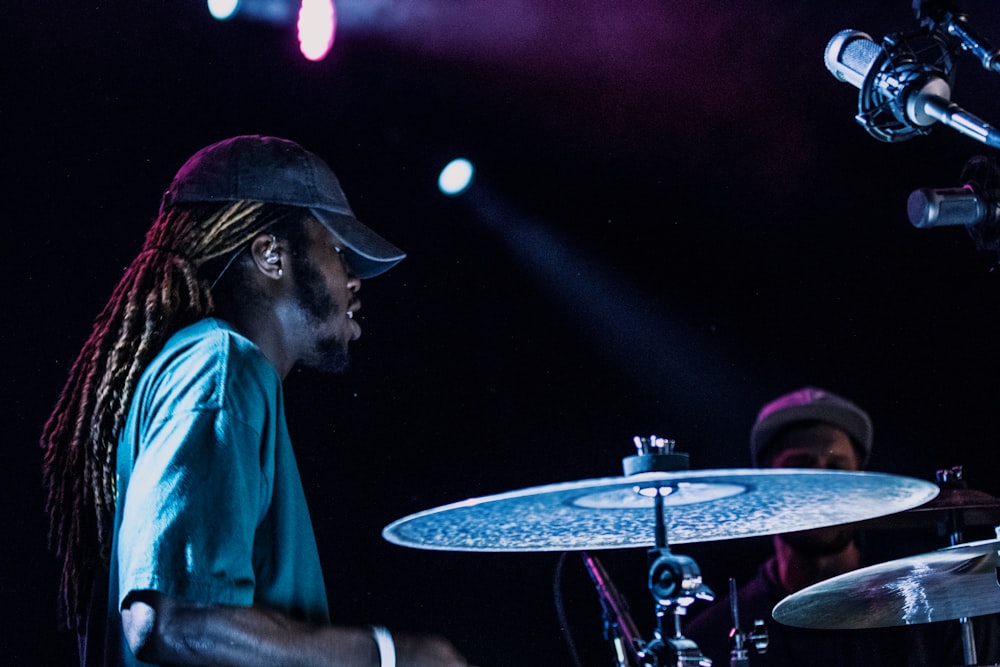 man playing drums beside man wearing gray fitted cap