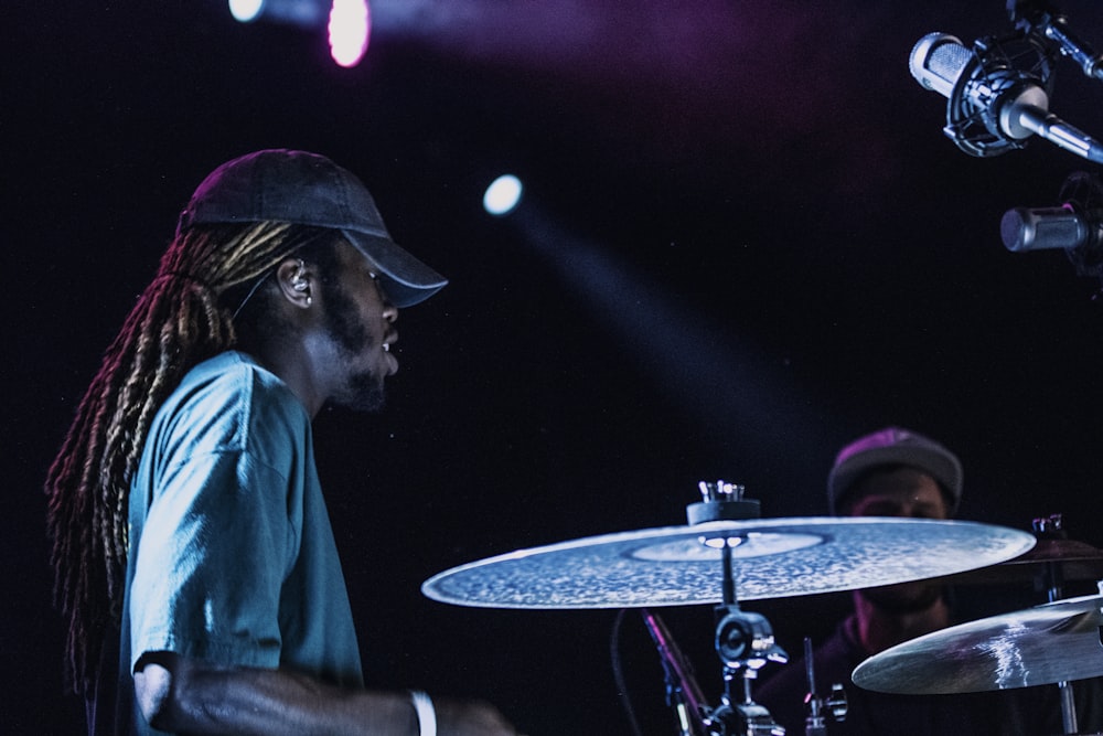 man playing drums beside man wearing gray fitted cap