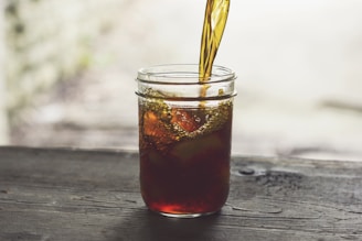 A jar is being filled with a dark liquid on a wooden surface, with light creating a soft, natural ambiance. The liquid forms bubbles and swirls as it is poured, adding a dynamic element to the scene.