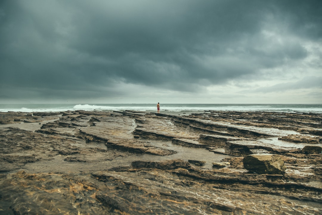 person in red shirt standing on brown rock formation near sea under white clouds during daytime