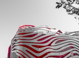 An architecturally striking building featuring smooth, wave-like patterns in red and white. The design is modern and innovative, with flowing lines that give a sense of movement. A bare tree's branches are visible in the top right corner against a pale sky.
