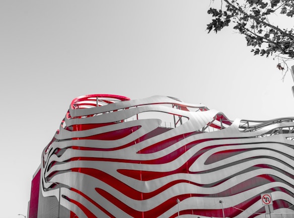 An architecturally striking building featuring smooth, wave-like patterns in red and white. The design is modern and innovative, with flowing lines that give a sense of movement. A bare tree's branches are visible in the top right corner against a pale sky.