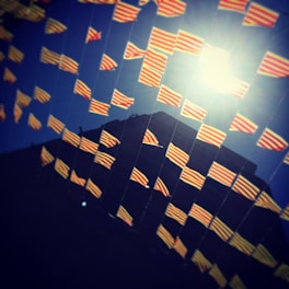 A series of small red and yellow striped flags are strung in rows, fluttering against a deep blue sky. The sunlight creates a bright spot, casting a glow that enhances the vibrant colors of the flags. A building silhouette is visible in the background.