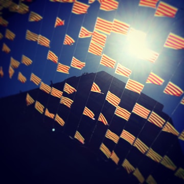 A series of small red and yellow striped flags are strung in rows, fluttering against a deep blue sky. The sunlight creates a bright spot, casting a glow that enhances the vibrant colors of the flags. A building silhouette is visible in the background.