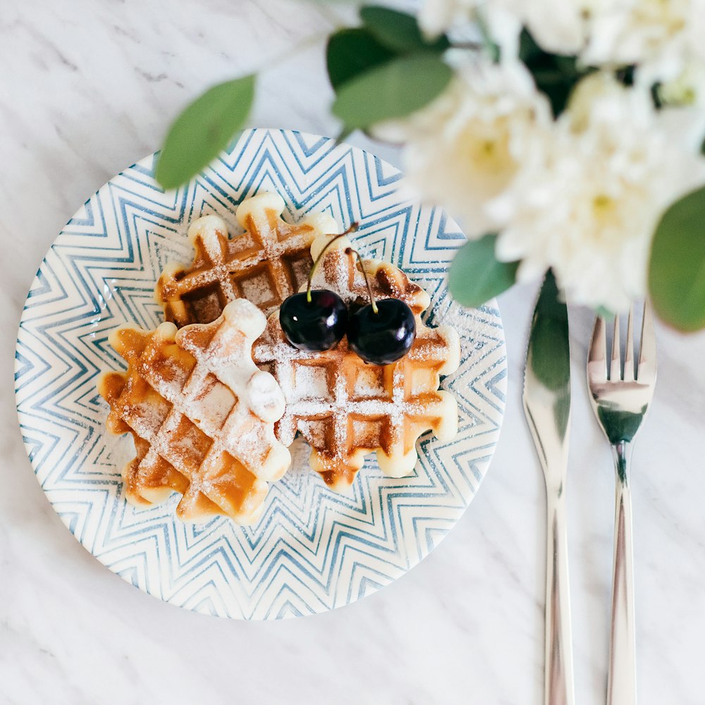 3 waffles on saucer with 2 cherry fruits beside fork and bread knife