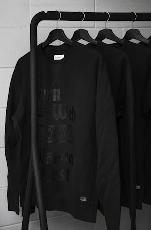 A row of black sweatshirts hanging on a black metal clothing rack against a light gray brick wall. The sweatshirts have a faint, stylized text on the front and appear to be identical in design and color. The overall setting gives a minimalist and modern impression.