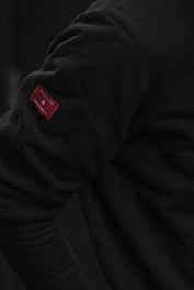 A close-up view of a person wearing a dark long-sleeve shirt. The focus is on the arm, highlighting a small red fabric tag with visible text.