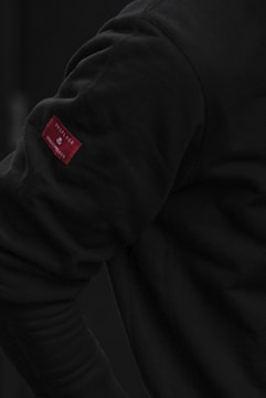 A close-up view of a person wearing a dark long-sleeve shirt. The focus is on the arm, highlighting a small red fabric tag with visible text.