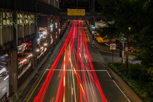 Long exposure photograph of a busy urban street, capturing streaks of red and white lights from moving vehicles in the night. The scene includes static traffic on one side and blurred light trails on a multi-lane road surrounded by greenery and metal structures. Various street signs and urban infrastructure elements such as bridges and overpasses add depth.