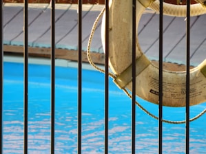 A lifebuoy hangs on a railing next to a swimming pool with clear blue water. Sunlight casts gentle shadows on the scene, and a wooden deck is visible in the background, just above the pool's edge.