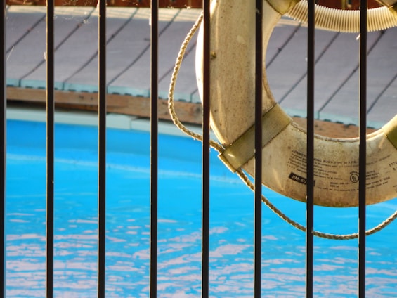 A lifebuoy hangs on a railing next to a swimming pool with clear blue water. Sunlight casts gentle shadows on the scene, and a wooden deck is visible in the background, just above the pool's edge.