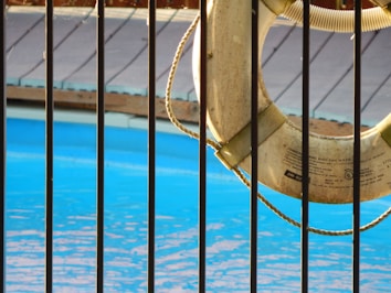 A lifebuoy hangs on a railing next to a swimming pool with clear blue water. Sunlight casts gentle shadows on the scene, and a wooden deck is visible in the background, just above the pool's edge.