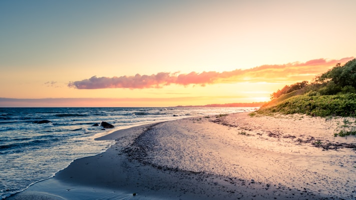 A tranquil beach with soft waves gently lapping against the shore, bathed in the golden light of a setting sun. The sky is a gradient of warm hues ranging from soft pinks and oranges near the horizon to pale blues higher up. A few scattered clouds add texture to the sky. Lush green vegetation grows along the right side, while the sandy beach curves gracefully towards the water.