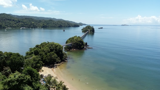 A serene coastal landscape featuring a long bridge connecting small lush islands surrounded by a tranquil blue sea. A sandy beach with gentle waves is visible, bordered by dense greenery and palm trees. The sky is clear with a few scattered clouds, and distant landmasses can be seen on the horizon.