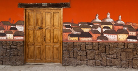 A rustic wooden door is set into a vividly painted wall that depicts an artistic mural of a village with several buildings topped by domes and pitched roofs. The wall features an upper section in bright orange and a lower section with a texture resembling stone bricks.