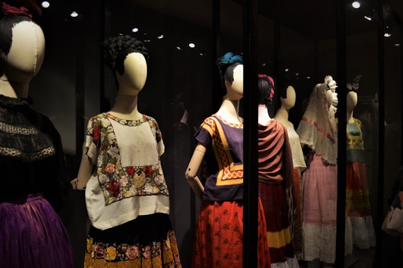 A collection of mannequins adorned with traditional, colorful dresses, featuring elaborate patterns and textiles. Each mannequin has a distinctive hairstyle and pose, creating a sense of cultural heritage and artistic expression.