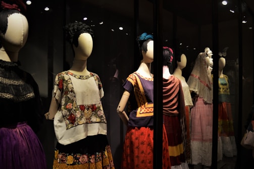 A collection of mannequins adorned with traditional, colorful dresses, featuring elaborate patterns and textiles. Each mannequin has a distinctive hairstyle and pose, creating a sense of cultural heritage and artistic expression.