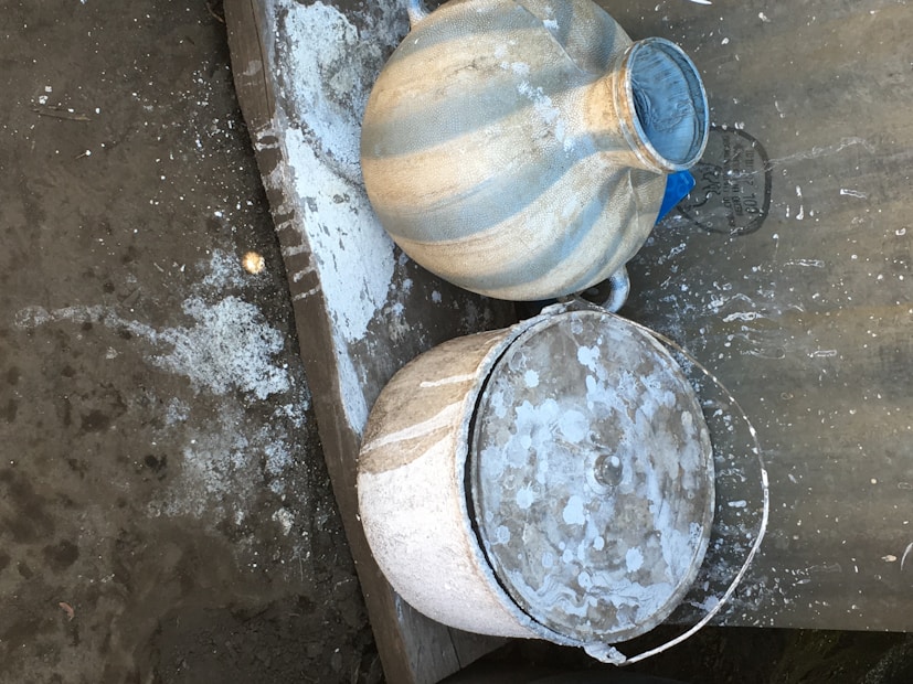 Two weathered clay pots are placed on a rough, uneven surface with scattered white powdery residue around them. One pot is larger with a narrow neck, while the other has a broader opening with a lid partially covering it. The materials and texture suggest they have been used for some time.