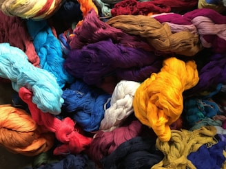 A vibrant assortment of dyed yarns is piled together, featuring a variety of colors including bright yellows, reds, blues, purples, and more.