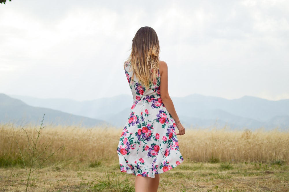 흰색, 보라색, 분홍색 꽃무늬 드레스를 입은 여자가 낮에 갈색 잎 잔디밭 근처에 서 있습니다.