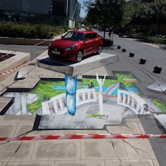 A red car is parked on a concrete platform that appears to be floating above an artistic 3D street mural. The mural creates an illusion of a grand architectural structure with white columns and green landscape. The scene is cordoned off with red and white striped tape.