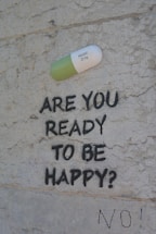 A large painted capsule labeled 'PROZAC 20 mg' is depicted on a textured wall. Below the capsule, the text 'ARE YOU READY TO BE HAPPY?' is spray-painted in black. Beneath this question, the word 'NO!' is also written in black.