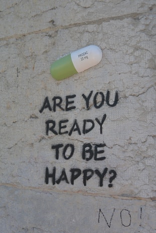 A large painted capsule labeled 'PROZAC 20 mg' is depicted on a textured wall. Below the capsule, the text 'ARE YOU READY TO BE HAPPY?' is spray-painted in black. Beneath this question, the word 'NO!' is also written in black.
