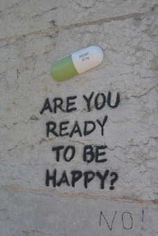 A large painted capsule labeled 'PROZAC 20 mg' is depicted on a textured wall. Below the capsule, the text 'ARE YOU READY TO BE HAPPY?' is spray-painted in black. Beneath this question, the word 'NO!' is also written in black.