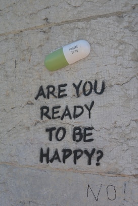 A large painted capsule labeled 'PROZAC 20 mg' is depicted on a textured wall. Below the capsule, the text 'ARE YOU READY TO BE HAPPY?' is spray-painted in black. Beneath this question, the word 'NO!' is also written in black.