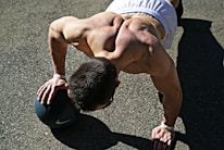 A muscular individual performing a push-up using a medicine ball on an outdoor surface, with sunlight casting strong shadows. The person is wearing athletic shorts and appears focused on their exercise.