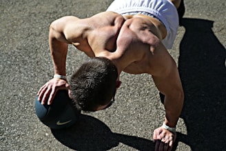 A muscular individual performing a push-up using a medicine ball on an outdoor surface, with sunlight casting strong shadows. The person is wearing athletic shorts and appears focused on their exercise.