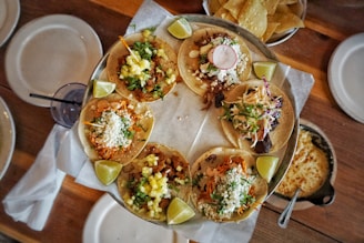A round metal tray holds a variety of colorful tacos garnished with fresh ingredients like cilantro, cheese, pineapple, radish slices, and lime wedges. Surrounding the tray are several white plates, a bowl of tortilla chips, a drink with a straw, and a skillet with melted cheese, all set on a wooden table.
