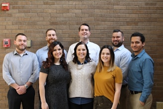 A group of eight people are standing in front of a brick wall, smiling at the camera. They are casually dressed and positioned closely together, suggesting a friendly and professional atmosphere. A fire alarm is visible on the wall to the left.